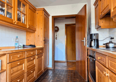 Küche mit Holzmöbeln, braunen Bodenfliesen und Blick in den angrenzenden Flur – funktionaler und gepflegter Kochbereich